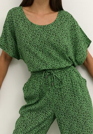 Femme portant un haut vert à manches courtes et un pantalon assorti avec un petit motif floral noir et une taille à cordon.