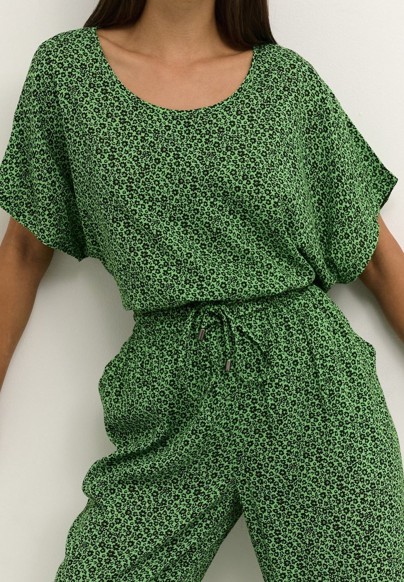 Femme portant un haut vert à manches courtes et un pantalon assorti avec un petit motif floral noir et une taille à cordon.