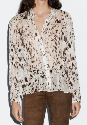 Femme portant un chemisier semi-transparent à manches longues avec des boutons, à motif tacheté marron et blanc, et un pantalon marron.