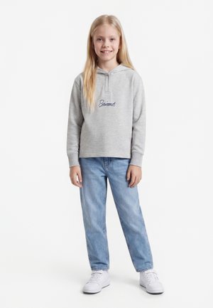 Jong meisje staat glimlachend, draagt een lichtgrijze hoodie met tekst, blauwe spijkerbroek en witte sneakers tegen een witte achtergrond.