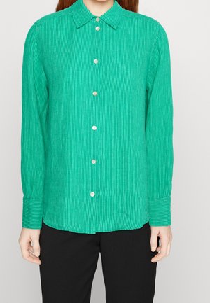 Femme portant une chemise verte à manches longues avec de fines rayures blanches verticales et un pantalon noir, debout les bras détendus.