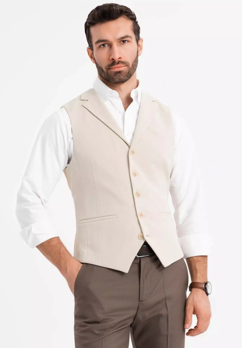 Beige Weste mit taillierter Passform, einreiher Design, strukturiertem Stoff und vier Knöpfen; getragen über einem weißen Hemd und braunen Hosen.