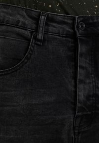 Vaqueros de mezclilla negra con una textura suave, que cuentan con un bolsillo delantero, cierre de botón y trabillas para cinturón. Se aprecia un brillo sutil en la tela.