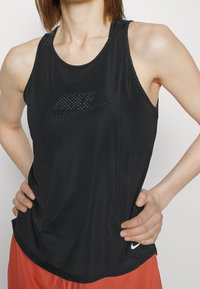 Czarny sportowy top na ramiączkach z teksturowanym wzorem, z perforowanym logo, okrągłym dekoltem i luźnym krojem. W połączeniu z pomarańczowymi szortami.