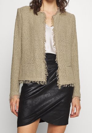 Vrouw draagt een beige gestructureerde open jas met franjes over een witte top en een zwarte rok van namaakleer met overslag.