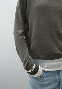 Maglione grigio a maglia con scollo a girocollo, tessuto testurizzato e orlo a coste, indossato sopra una camicia bianca a maniche lunghe, abbinato a jeans in denim blu.
