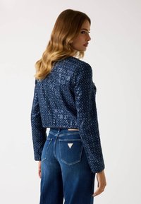 Donna con capelli biondi ondulati che indossa una giacca lunga a maniche lunghe blu navy con paillettes e jeans scuri a vita alta, di profilo.