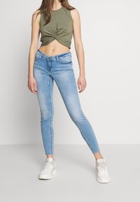 Jeans de mezclilla azul delgados con un lavado desgastado, que presentan un diseño clásico de cinco bolsillos. Combinados con una camiseta sin mangas verde olivo recortada.