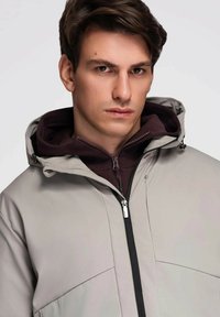 Jasnoszara kurtka soft-shell z zamkiem błyskawicznym, regulowanym kapturem i nakładanym ciemnofioletowym wewnętrznym kapturem. Charakteryzuje się nowoczesnym, schludnym wzornictwem.