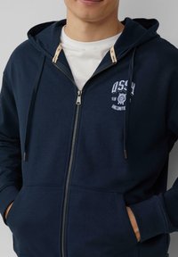 Felpa con zip color navy, dotata di tasca a marsupio, polsini a costine e una grafica decorativa sul petto. Presenta zip color crema.