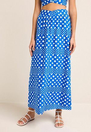 Femme portant une jupe longue à motifs géométriques bleus et un haut court assorti, avec des sandales blanches à brides, debout devant un fond uni.