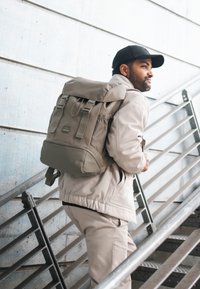 Vihreä kangasreppu, jossa on rullauskansilukko, säädettävät hihnat ja metalliosat, jota henkilö kantaa portaikossa.