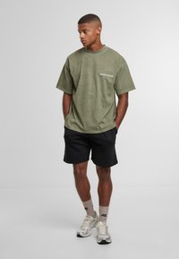 Mladý muž v olivově zeleném oversized tričku, černých šortkách, béžových ponožkách a světlých teniskách, stojící s rukama v kapsách.