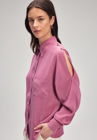 Camicia rosa chiaro a maniche lunghe con chiusura a bottoni, taschino sul petto e dettagli a taglio sulle spalle. Tessuto morbido e liscio.