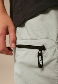Jasnoszare spodnie z boczną kieszenią zapinaną na zamek, z czarnym uchwytem zamka z napisem "SUBSKRYBUJ". Gładka faktura materiału i luźny krój.