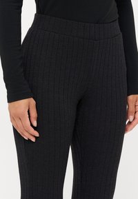 Fekete bordás leggings magas derékkal, puha anyagból, amely függőleges csíkokkal van díszítve. A közeli kép a kéz elhelyezkedését mutatja az anyagon.