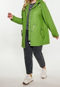 Vihreä vedenpitävä takki, jossa on istuva leikkaus, vetoketjutasku, kiristysnauha vyötäröllä ja kaksi sivutaskua. Malli käyttää raidallista yläosaa ja tummia farkkuja.