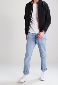 Man wearing a black zip-up jacket over a white t-shirt, light blue rolled-up jeans, and white sneakers standing against a plain background.