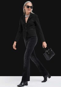 Schwarzer, strukturierten Blazer mit taillierter Passform, kombiniert mit dunklen Jeans mit Nietenakzenten. Accessoires sind eine kleine schwarze Handtasche und Sonnenbrille.