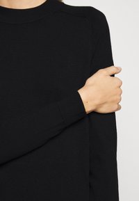 Personne portant un pull noir à manches longues avec des poignets côtelés, tenant son bras gauche au-dessus du coude avec la main droite, sur un fond uni.