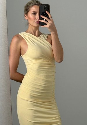 Donna con capelli biondi, vestito giallo pallido senza maniche con arricciature, che scatta un selfie allo specchio con uno smartphone nero davanti a una parete grigia uniforme.