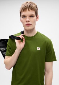 Zöld pamut póló kerek nyakkal, egy kis CK logós folttal. A modell az egyik vállán egy fekete táska pántot tart.