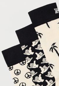 Τρία ζευγάρια ασπρόμαυρες κάλτσες με σύμβολα ειρήνης, χειρονομίες shaka και μοτίβα φοίνικα σε ανοιχτό φόντο.