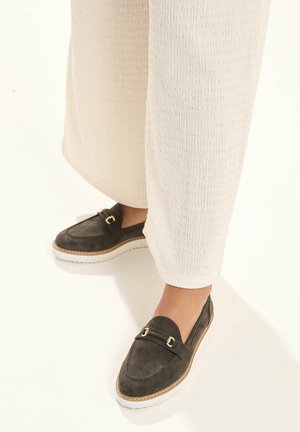 Voeten met donkergrijze suède loafers met gouden accenten en witte zolen, gecombineerd met gestructureerde off-white wijde broekspijpen.