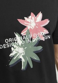 Camiseta negra con gráficos florales en rosa y verde y texto blanco que dice "ORIGINALS © 1990 DISEÑADA EN CPNHGN." Material de algodón suave.