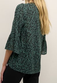 Blouse à motifs verts avec une surface texturée, arborant un design abstrait noir. Elle possède des manches évasées et une coupe décontractée.