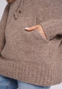 Brązowy dzianinowy sweter wykonany z miękkiej tkaniny, z przednią kieszenią i ściągaczowym dołem. Zbliżenie pokazuje fakturę i detale materiału.