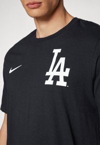 Black cotton t-shirt features a white "LA" logo and a small white Nike swoosh on the left chest area, with a classic crew neck design.