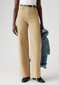 Pantalones de pana anchos color beige con cintura alta, que cuentan con bolsillos frontales, un cinturón negro y una chaqueta de mezclilla colgada de un brazo.