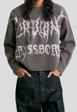 Persoon draagt een grijze trui met gestileerde witte tekst "KEY POINT" en "ABYSSBORN," handen in de zakken van een zwarte broek.