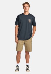T-shirt en coton bleu foncé avec une poche poitrine ornée d'un motif circulaire, associée à un short kaki et des baskets noires avec des chaussettes blanches.