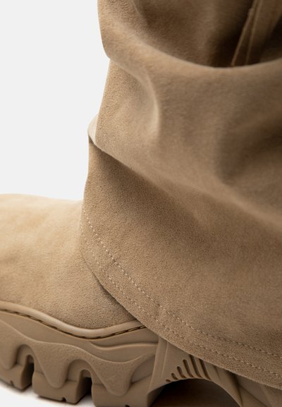 Κοντινή λήψη από μποτάκι σε μπεζ σουέντ με χαλαρή πάνω μεριά και χοντρή, ραβδωτή ελαστική σόλα.