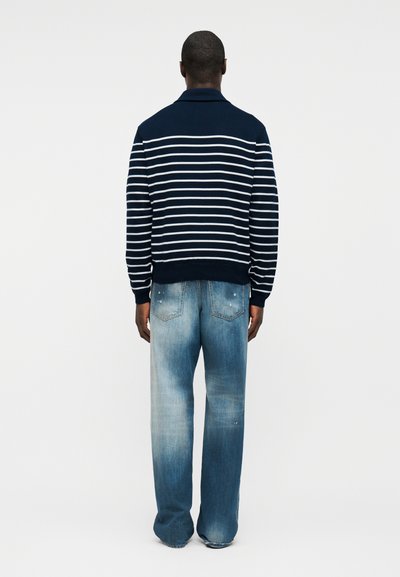 Homme debout, tourné de dos, portant un pull à rayures bleu marine et blanc ainsi qu'un jean bleu délavé avec des poches arrière.