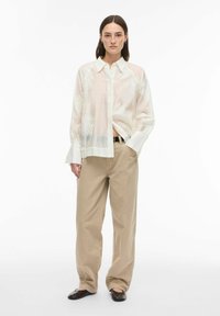 Blouse légère et transparente avec broderie florale, de couleur blanche, manches longues et col avec revers. Associée à un pantalon beige, ample, à jambes droites.