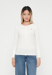 Biely dlhý rukáv henley tričko s okrúhlym výstrihom, gombíkovou légou a malým červeným logom. Materiál je mäkký a mierne priliehavý.