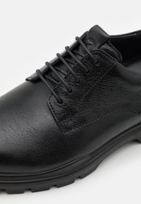 Chaussure en cuir noir à texture lisse, avec un bout arrondi et un laçage traditionnel. Semelle noire épaisse avec un motif robuste.