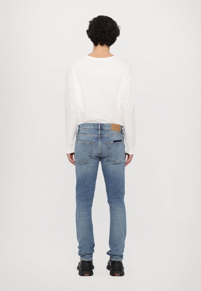 Lichtgewicht witte blouse met lange mouwen in combinatie met strakke blauwe denim jeans. De jeans hebben een bruine leren patch en twee achterzakken. Zwarte sneakers.