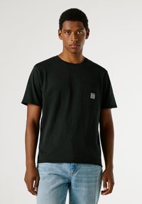 Μαύρο κοντομάνικο μπλουζάκι με τσέπη στο στήθος και ετικέτα, κατασκευασμένο από υφασμάτινη ύλη, φορεμένο με ανοιχτό μπλε τζιν παντελόνι.