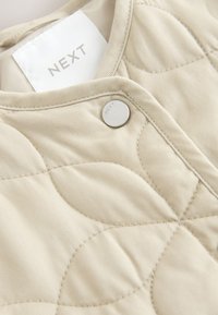 Beige quiltet stof med cirkulær syning, metal trykknap graveret "NEXT" og hvidt tøjmærke med mærket "NEXT".