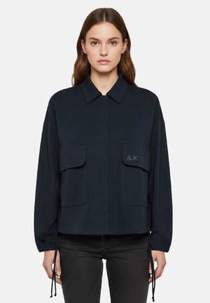 Giovane donna con capelli castani lisci indossa una giacca blu navy con tasche con patta sul davanti e polsini con coulisse, abbinata a jeans scuri.