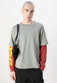 T-shirt en coton gris à manches courtes, avec un logo rouge. Longue manche rouge en dessous avec un imprimé graphique jaune. Détail d'un sac à épaule noir.