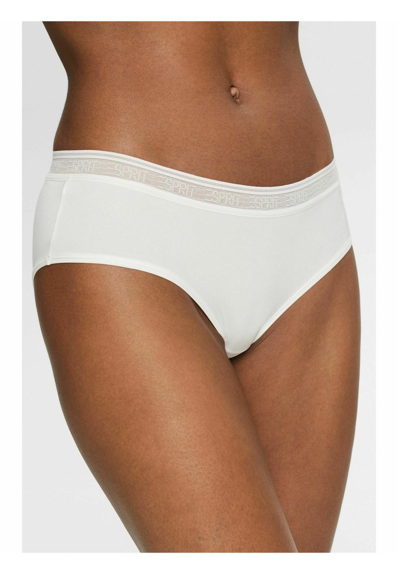 Frau, die weiße Hipster-Unterwäsche mit einem elastischen Bündchen mit Markenlogo trägt, steht vor einem schlichten Hintergrund.