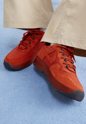 Personne portant un pantalon beige et des baskets Nike rouge vif, debout sur un sol recouvert d'un tapis bleu clair.
