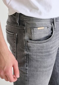 Jean en denim gris avec un design classique à cinq poches. Comprend des légères déchirures et une étiquette en métal sur la poche arrière. La texture est douce et lisse.