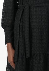 Abito nero con un motivo a foglia testurizzato, con vita cinchata e una cintura legata. Maniche lunghe con dettagli in pizzo intricati sui polsini.