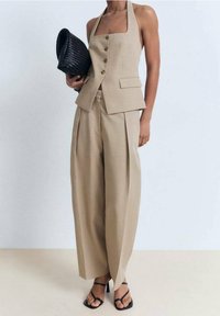 Beige, op maat gemaakt broekpak met een halternek-top, voorknopjes en licht gestructureerde stof. Gecombineerd met een geweven zwarte clutch en sandaaltjes met bandjes.